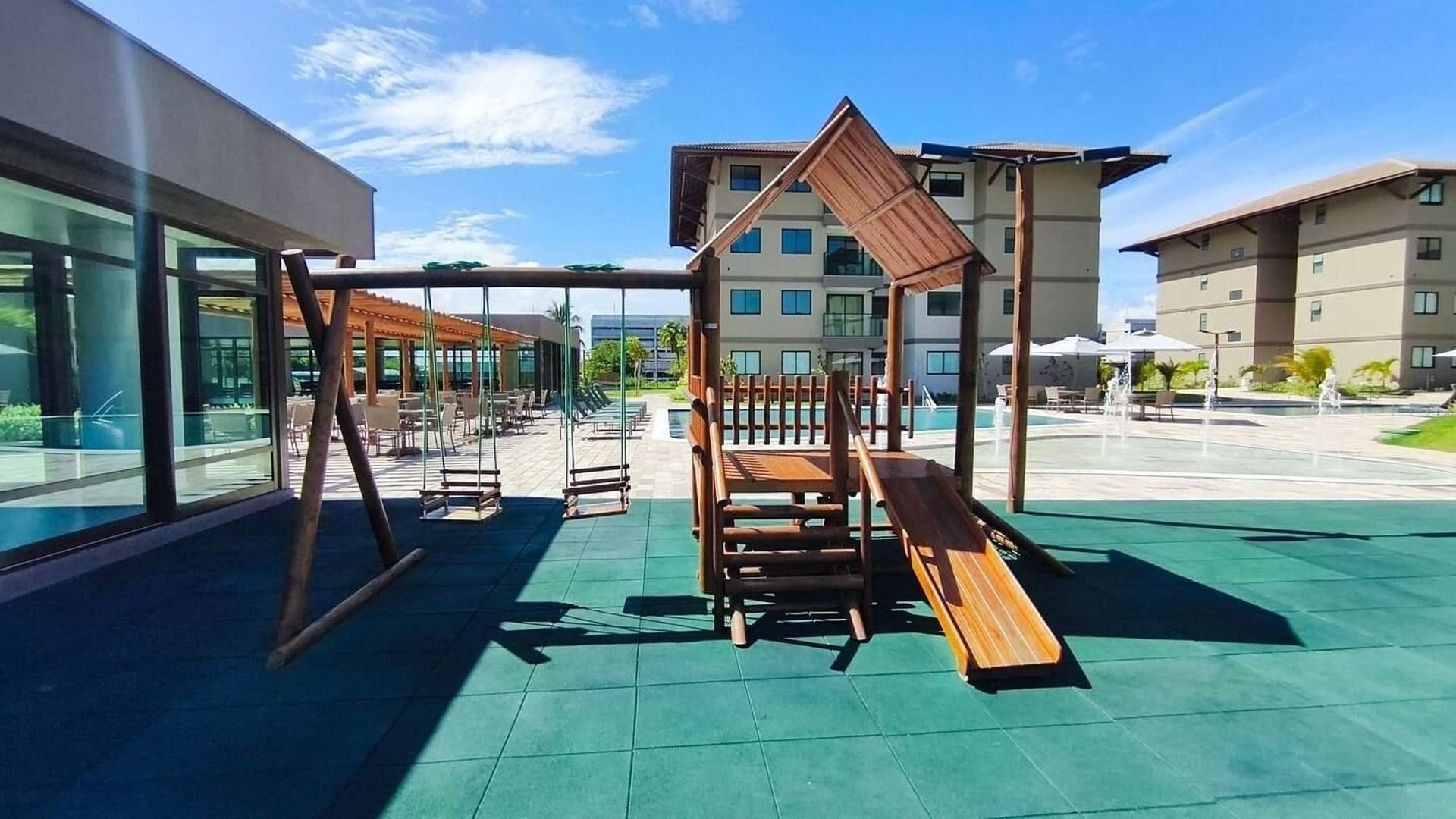 An outdoor play area with a wooden slide and swings, surrounded by buildings with large windows.