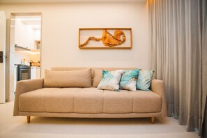 Un sofá beige con tres almohadas, una obra de arte de madera, y vistas a una cocina y comedor.