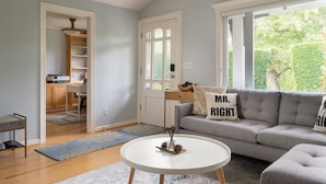 A living room with a grey sofa, a round white coffee table, and a view of a kitchen and garden through a large window. - Private Garden Home in Madison Valley (Seattle)