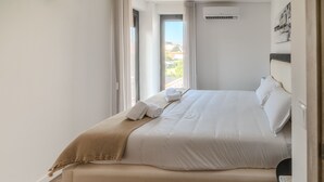 Una cama bien hecha con sábanas blancas y un plaid color beige, una mesita de noche con un control remoto y una ventana con cortinas.
