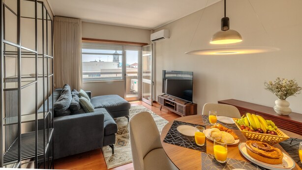 A modern living room with a sofa, television, dining table with breakfast, and a balcony with a view.