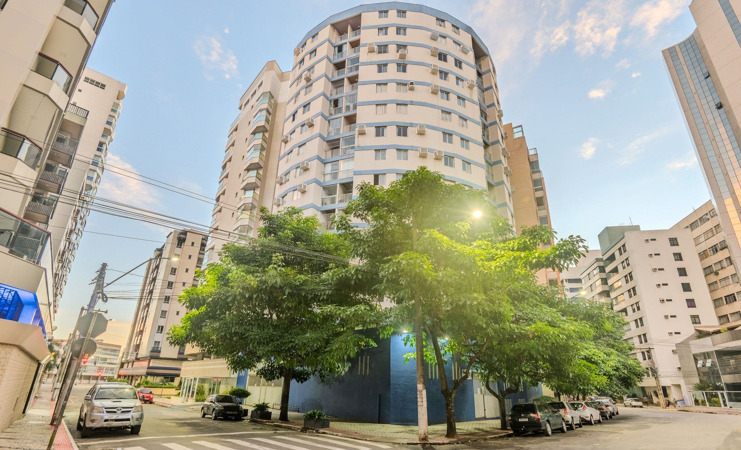 A multi-story residential building with balconies, a tree-lined street, and parked cars.