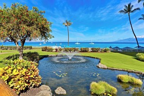 A landscaped garden with a pond, palm trees, and a view of the ocean with sailboats.