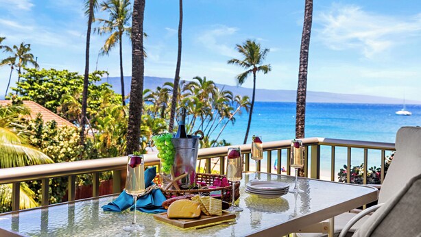 Ein Tisch mit einer Flasche, Gläsern und Speisen auf einem Balkon mit Blick auf einen Strand, an dem Palmen und das Meer zu sehen sind.