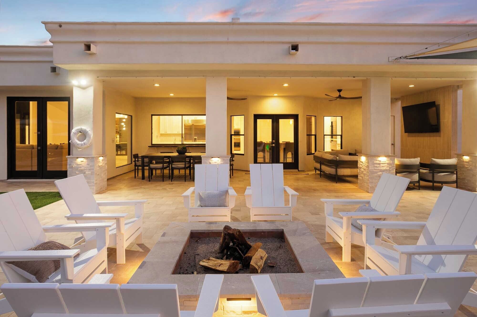A covered outdoor patio with white lounge chairs, a fire pit, and a dining area with a table and chairs.