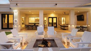 A covered outdoor patio with white lounge chairs, a fire pit, and a dining area with a table and chairs.