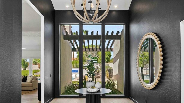 Un interior moderno con una pared oscura, una mesa redonda con adornos, un espejo grande y vistas a un espacio exterior con plantas y una pérgola.