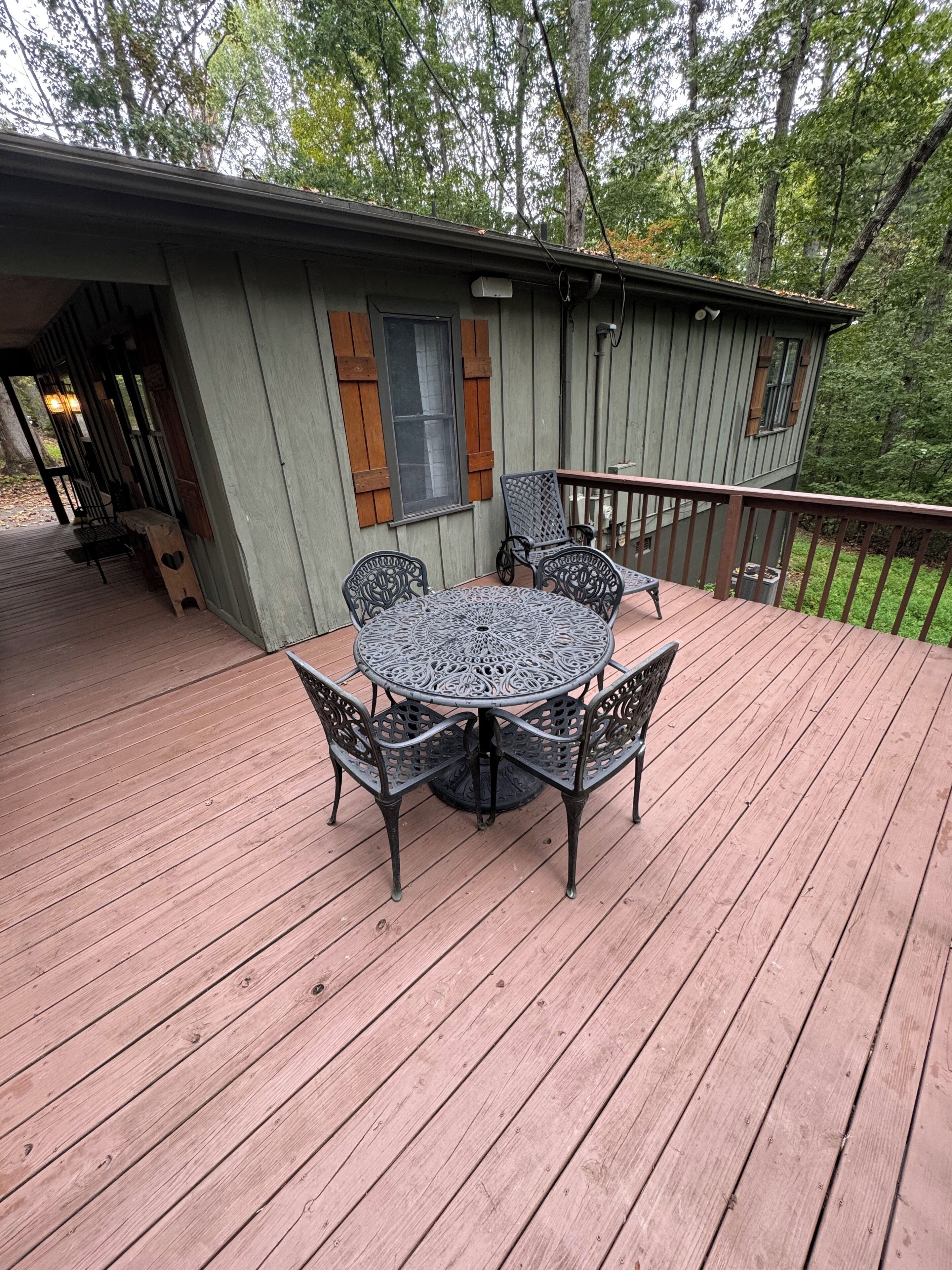 A wooden deck with a round table and chairs, a small porch area, and a building with shutters and a window.