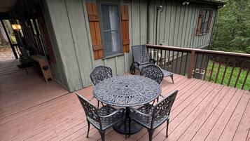 A wooden deck with a round table and chairs, a small porch area, and a building with shutters and a window.