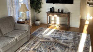 Una sala de estar con un sofá gris, un tapete estampado, una mesita de madera con una lámpara, una planta en maceta y un televisor de pantalla plana montado en una pared blanca con paneles.