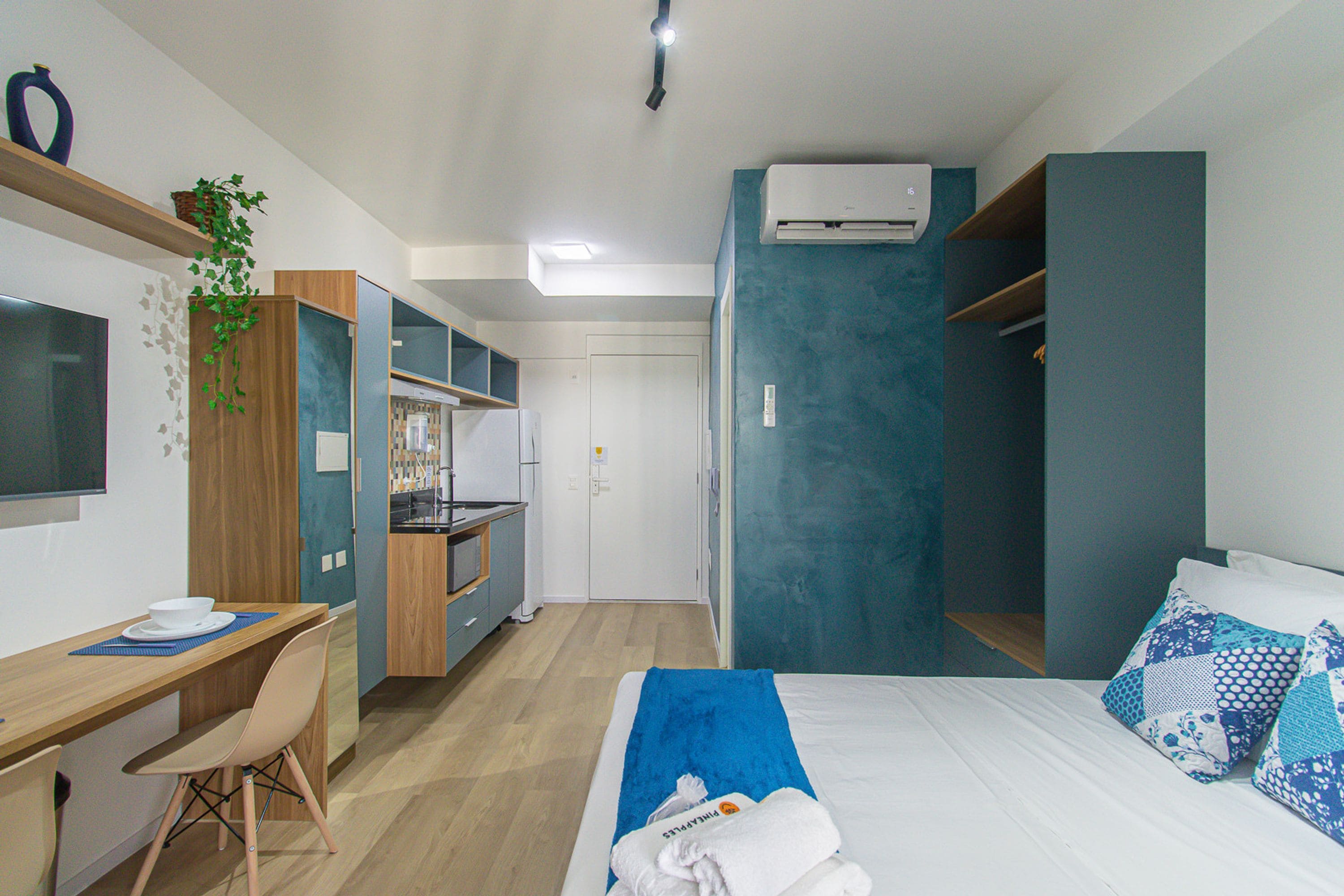 Ein gemütliches Hotelzimmer mit Küchenzeile, einem Bett mit blauer Bettwäsche, einem Schreibtisch mit Stuhl und einer kleinen Pflanze.