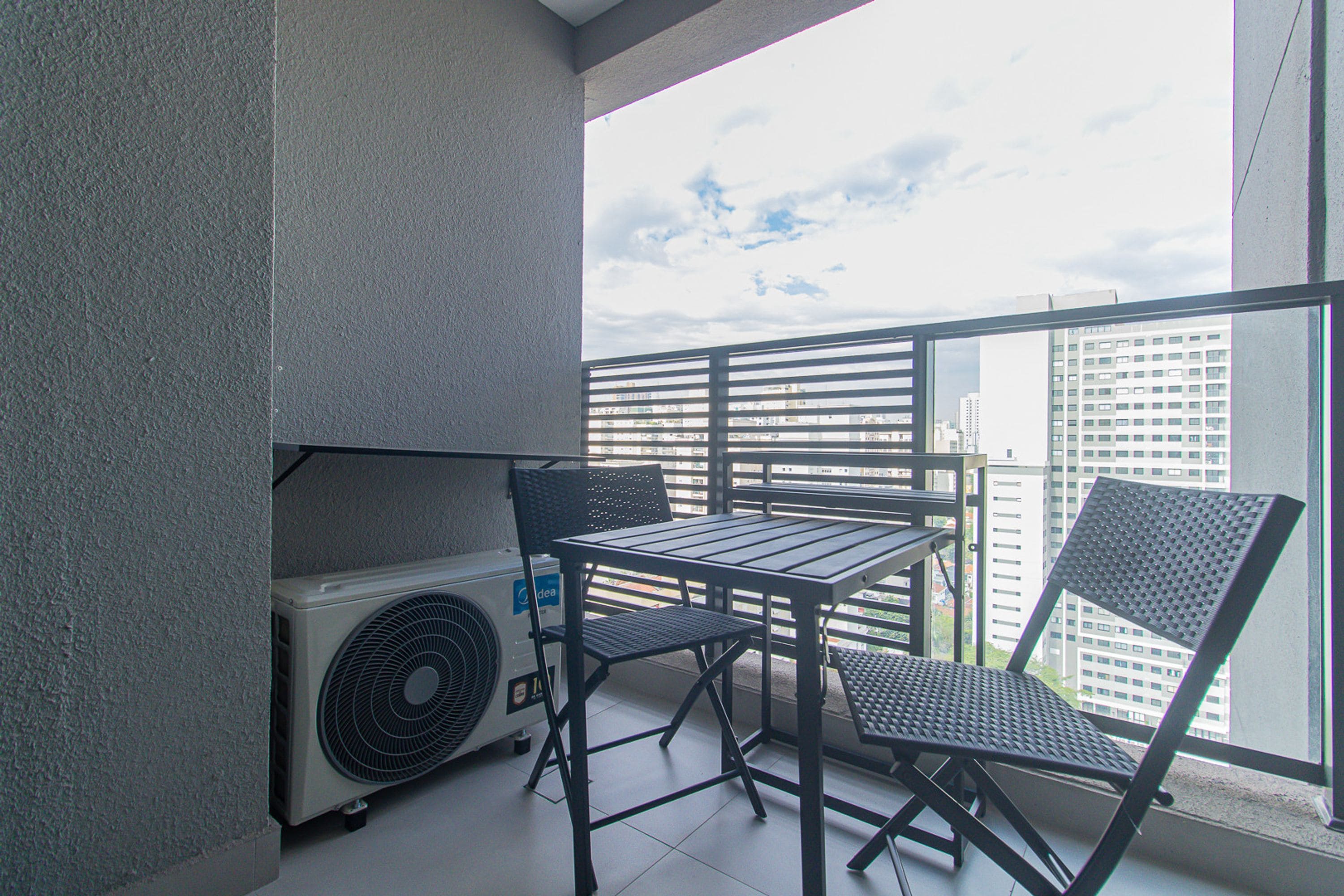 A balcony with a table and chairs, an air conditioning unit, and a view of city buildings.