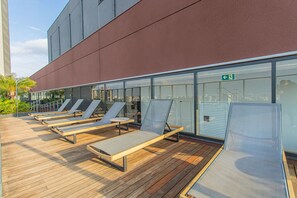 A rooftop lounge area with sun loungers and a glass wall overlooking the city.