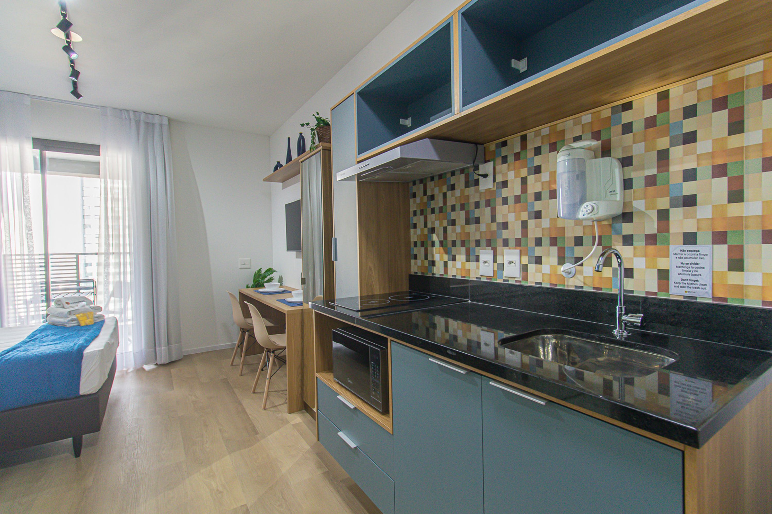 A modern kitchen with a black countertop, a built-in oven, and a microwave. There is a dining area with a table and chairs, and a bed with a blue blanket.