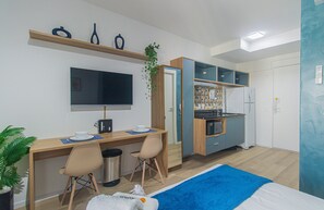 A compact hotel room with a wooden desk, two chairs, a bed with a blue blanket, a TV, a small kitchenette, and a potted plant.