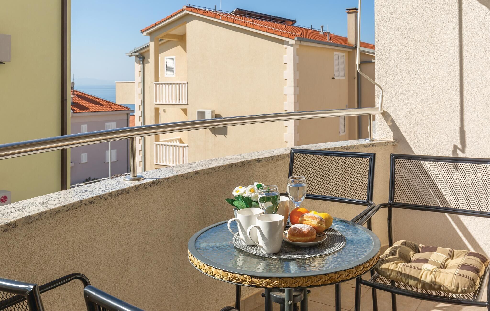 Ein Balkon mit rundem Tisch, zwei Stühlen, einem kleinen Teppich, einem Kissen, einer Blumenvase, einem Teller mit Gebäck und zwei Gläsern Wasser.