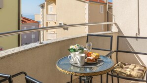 Un balcón con una mesa redonda, dos sillas, un tapete pequeño, un cojín, un jarrón con flores, un plato con pasteles y dos vasos de agua.