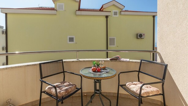 Un balcon avec deux chaises et une petite table, un sol carrelé et, en arrière-plan, un bâtiment avec un toit rouge.
