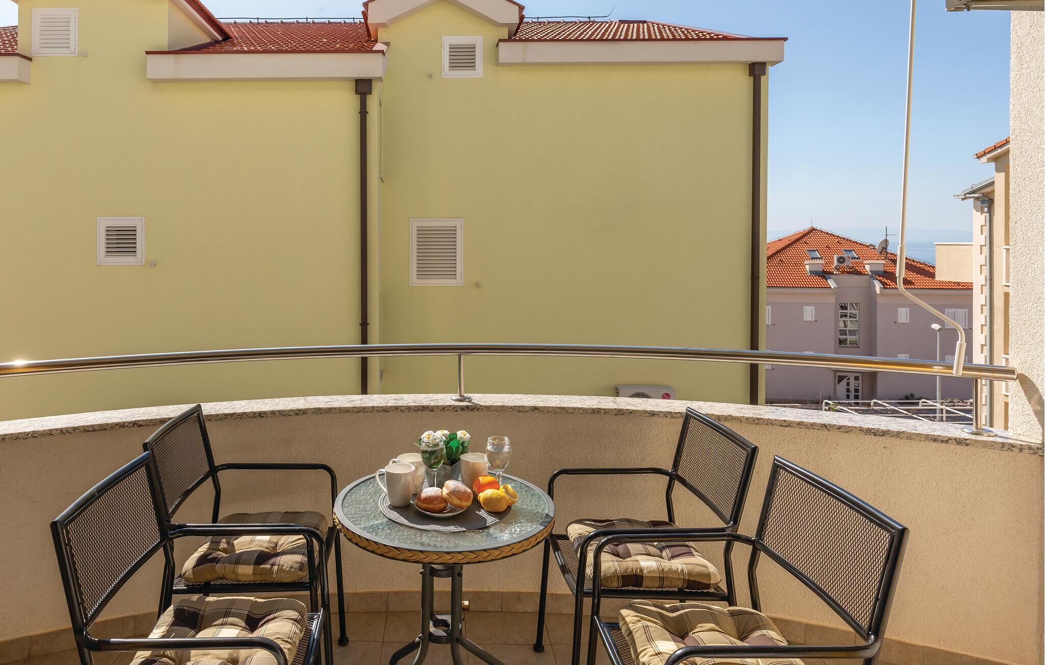 En balkon med et rundt bord og fire stole, et lille bord med et glas vand, en kop og et fad med kager.