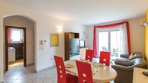 Un comedor con una mesa redonda, sillas rojas y un sofá. Hay una zona de cocina con un refrigerador y armarios, y un baño con inodoro y lavamanos.