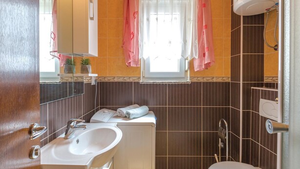 A bathroom with a sink, mirror, window with curtains, and a shower area.