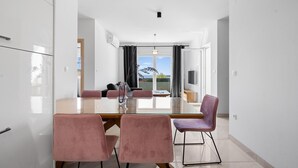 Un espace repas moderne avec une table en verre, des chaises roses et une vue sur l’extérieur grâce à de grandes fenêtres.