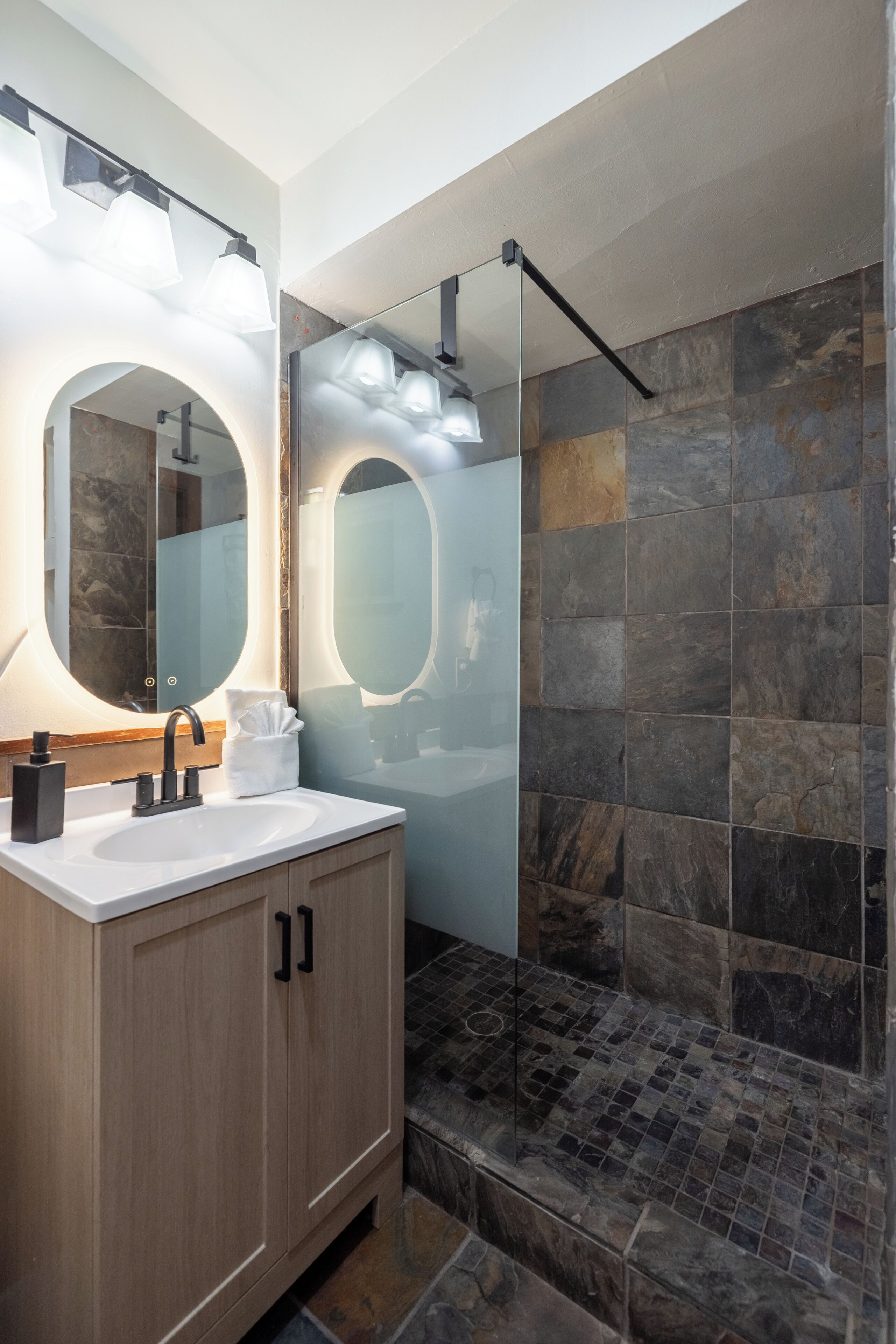 A modern bathroom with a large mirror, a sink, and a shower area with stone tiles.