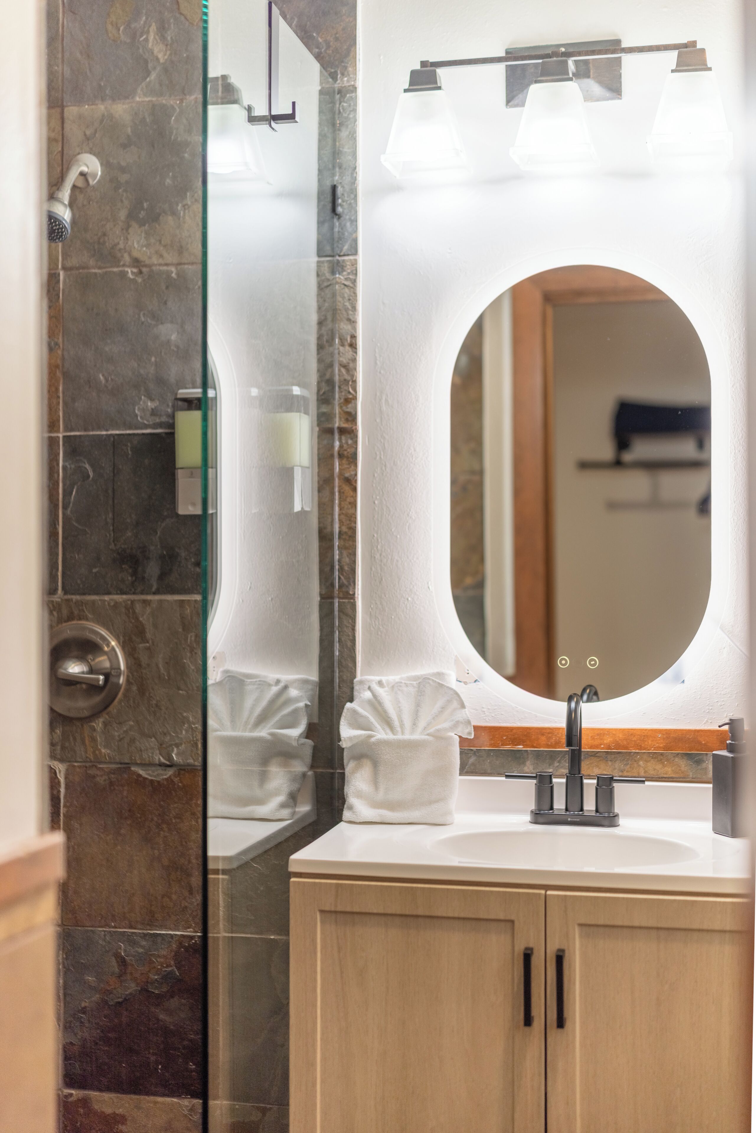 Une salle de bain avec un mur en marbre, une douche en verre, un lavabo blanc doté d’un robinet moderne et un grand miroir.