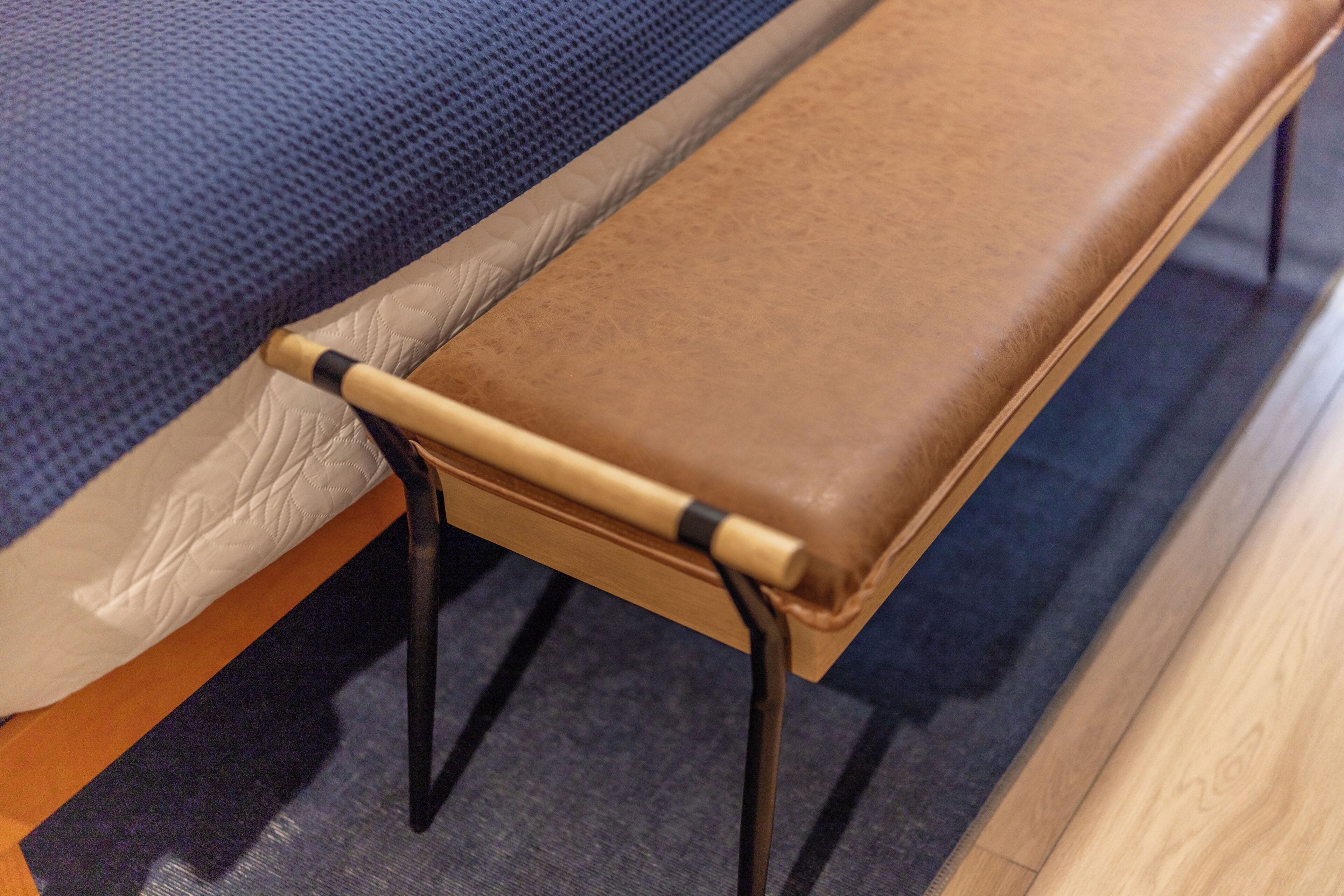 Una cama con colchón azul texturizado, un banco de cuero marrón y patas de metal negro.