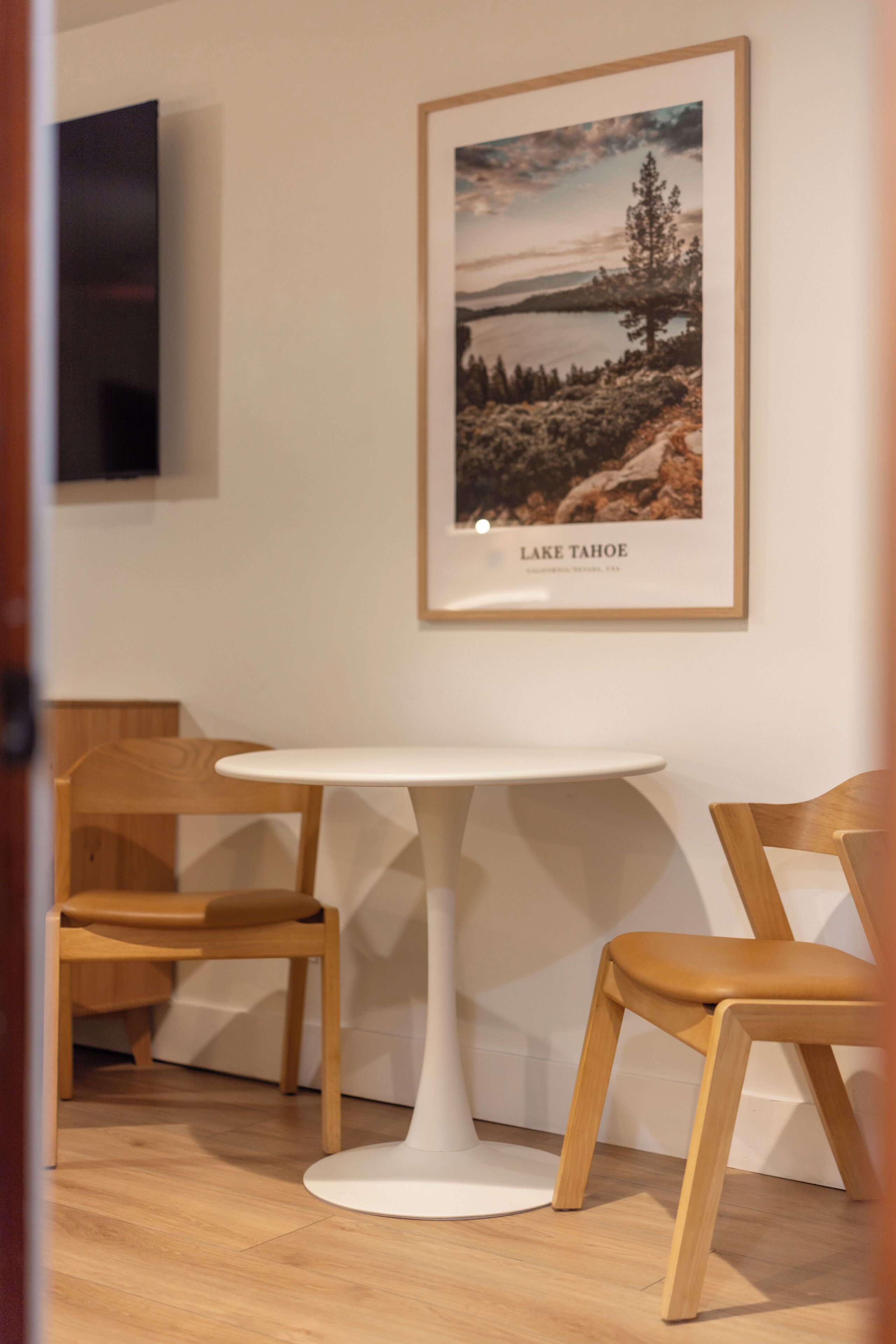 A room with a white table and two wooden chairs, a framed picture of Lake Tahoe on the wall, and a television mounted on the wall.