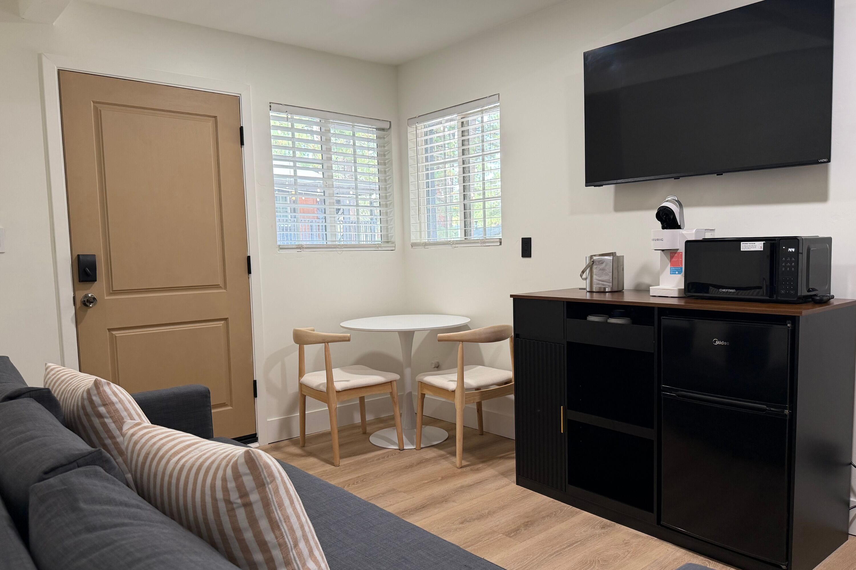 A compact living space with a sofa, a small dining table with two chairs, a kitchenette with a microwave, and a TV mounted on the wall.