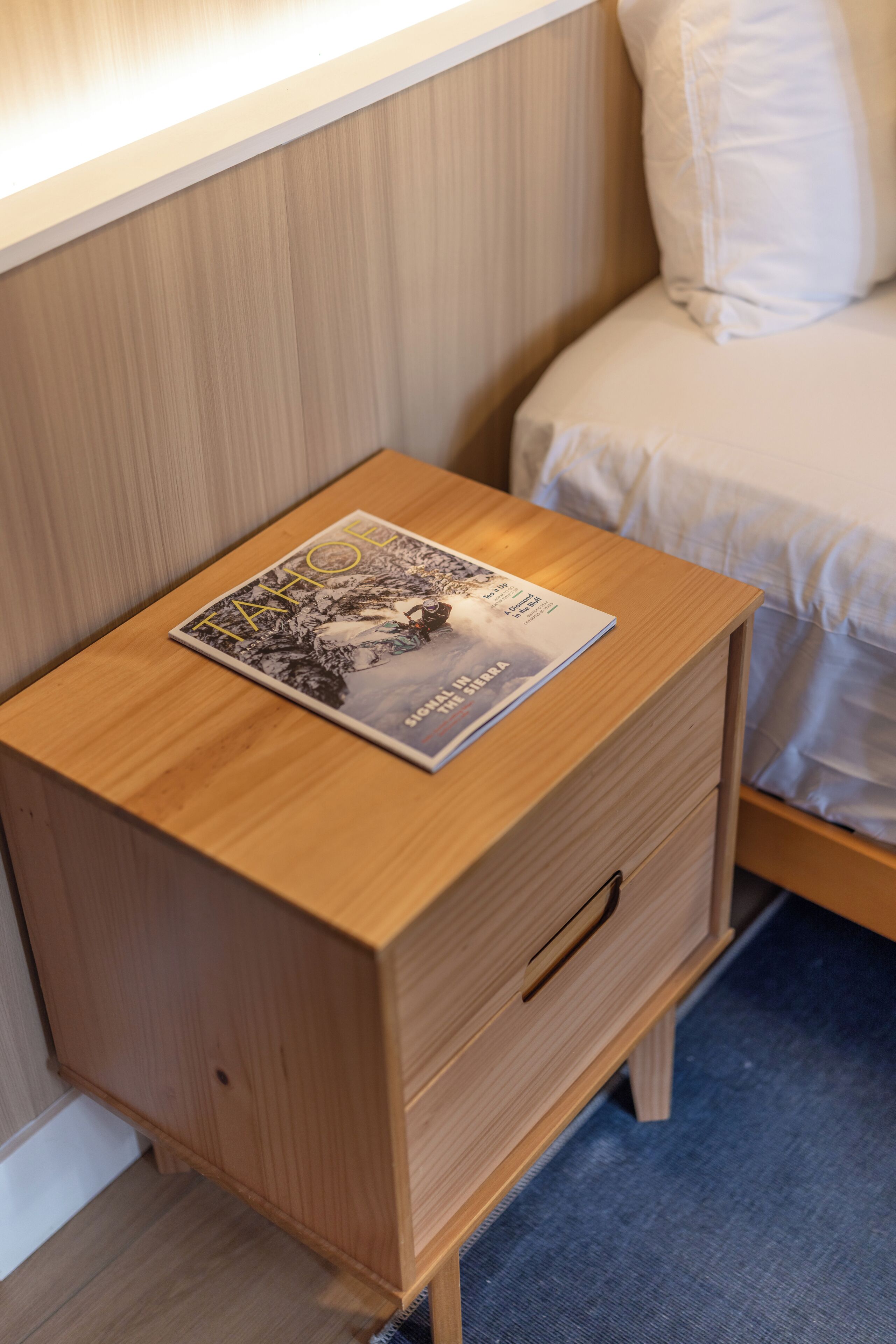 Une table de chevet en bois, avec un magazine posé dessus, un tiroir, et un lit avec une literie blanche en arrière-plan.