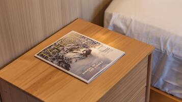 Une table de chevet en bois, avec un magazine posé dessus, un tiroir, et un lit avec une literie blanche en arrière-plan.
