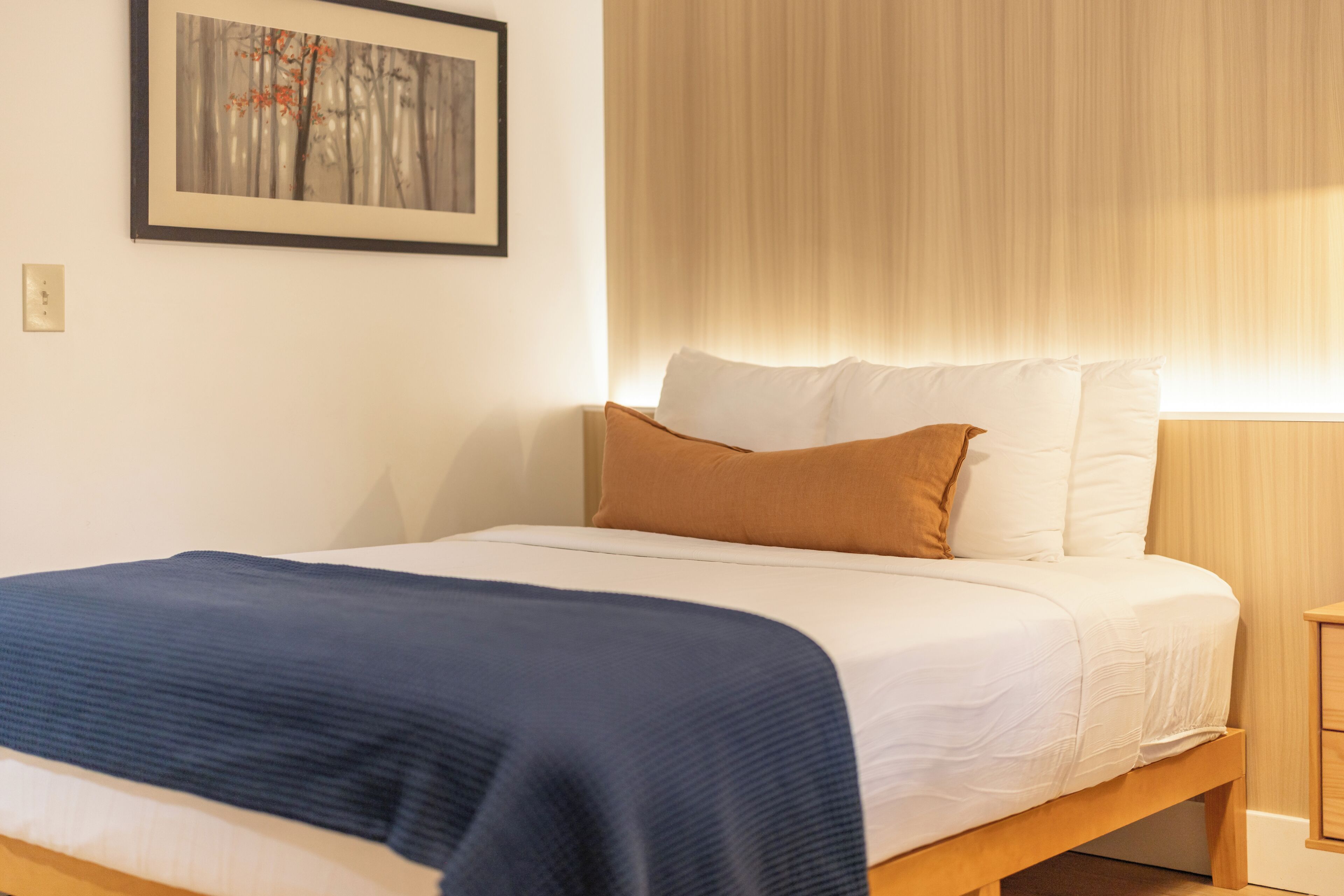 Una cama bien hecha con una colcha azul, almohadas blancas y un cojín decorativo marrón.