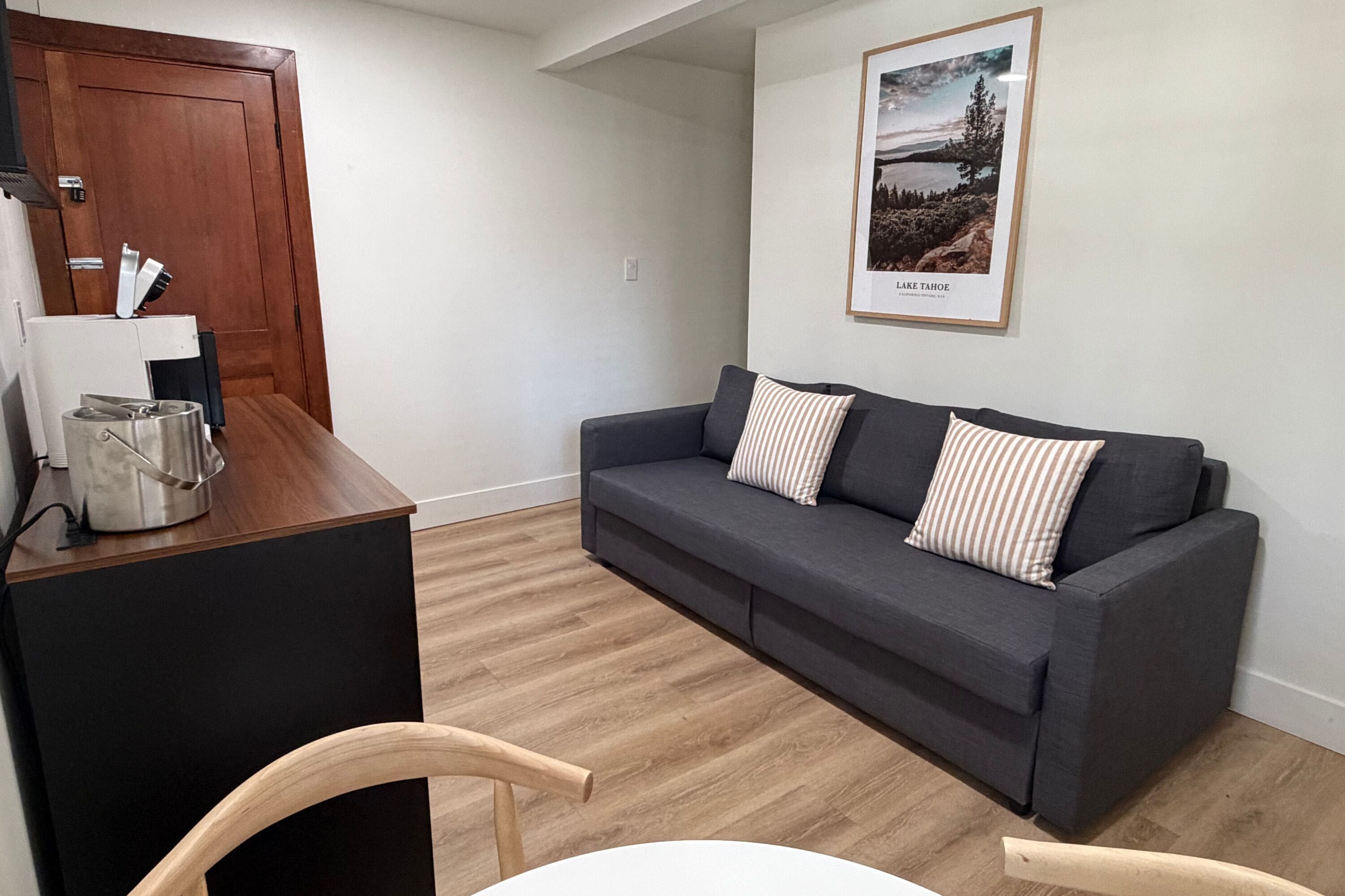 Un salón con un sofá gris oscuro, dos almohadas a rayas, una mesa de centro de madera y un cuadro enmarcado de un paisaje en la pared.