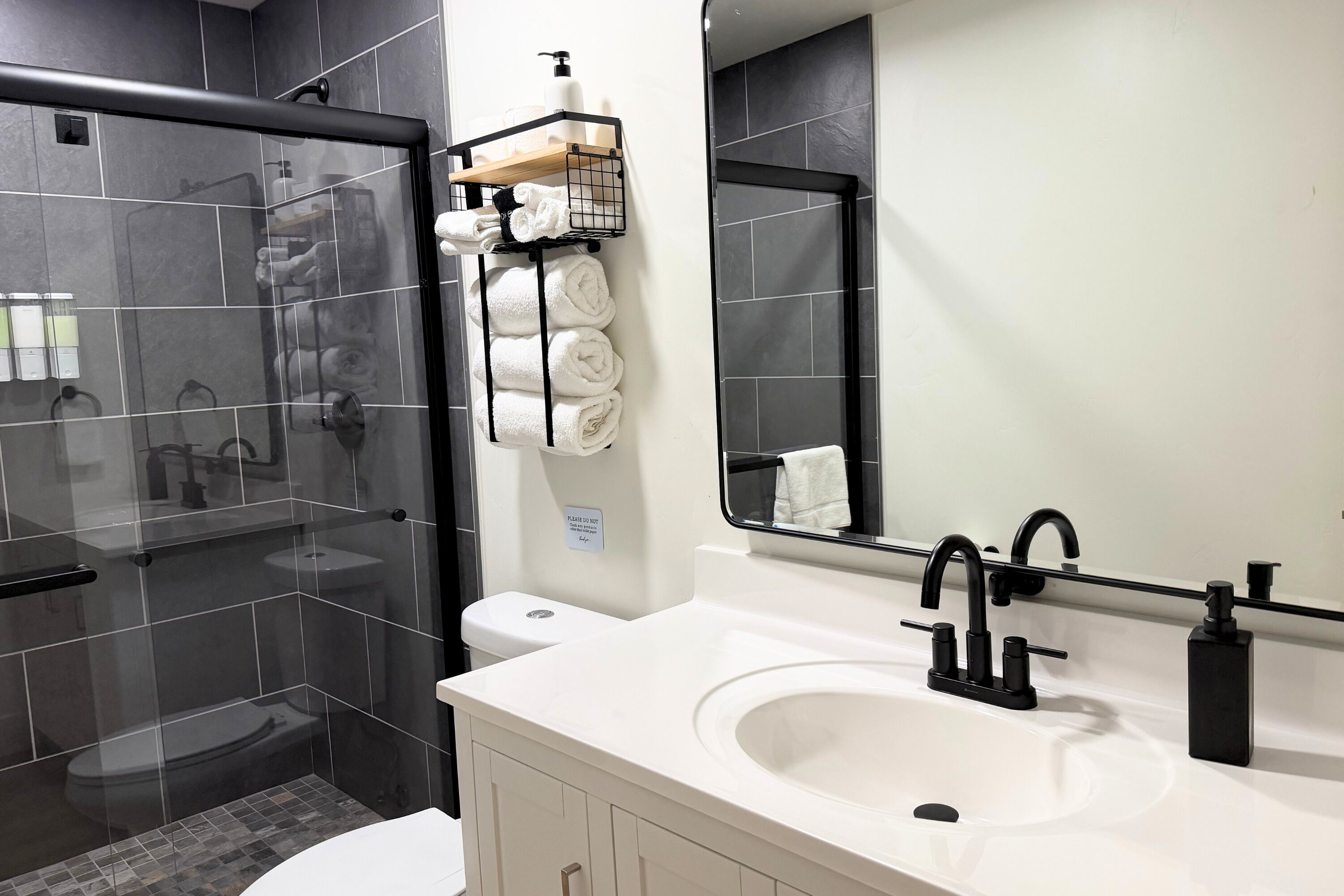 Un baño moderno con cabina de ducha de vidrio, lavamanos blanco con grifería negra y un perchero con toallas dobladas con cuidado.