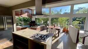 Una cocina moderna con estufa a gas, horno empotrado y una isla grande. Hay un comedor con mesa y sillas, y un balcón con vistas a árboles y pájaros.