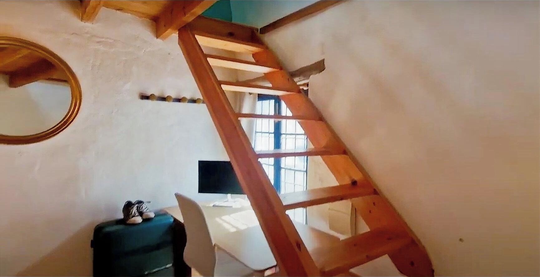 A room with a wooden ladder, a desk, a chair, a small table, and a window.