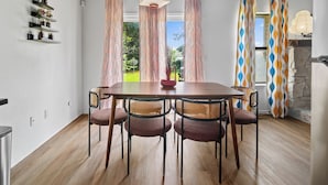 A dining area with a wooden table and four chairs, a hanging light fixture, and a view of the outdoors through patterned curtains. - Groovy Getaway - Retro Vibes and Modern Comfort (Tulsa)