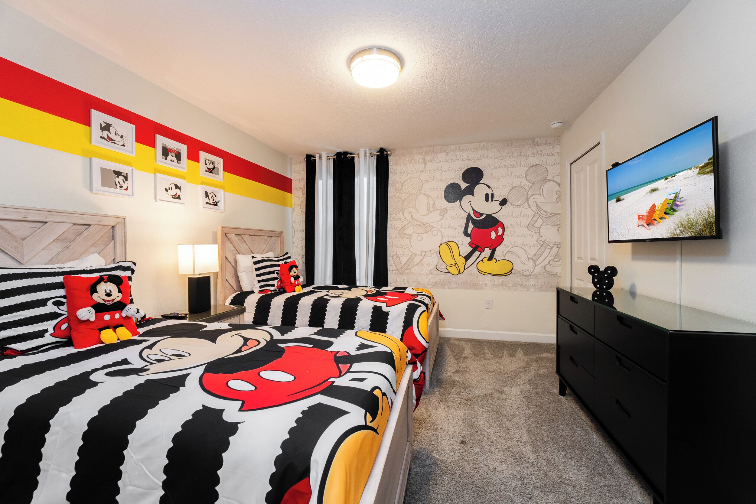 Une chambre avec deux lits, chacun agrémenté de linge de lit aux motifs de Mickey Mouse, une fresque murale représentant Mickey Mouse, un téléviseur à écran plat fixé au mur et une commode.