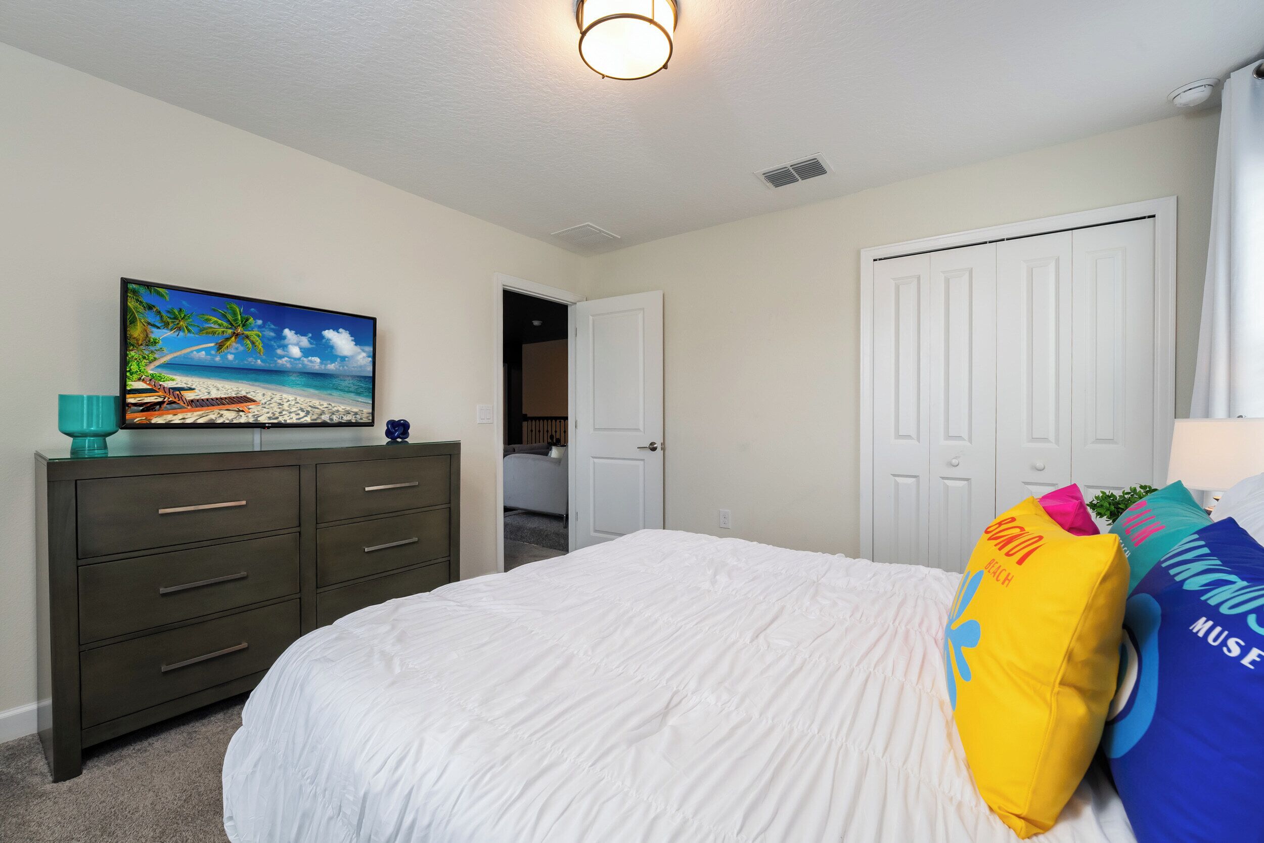 A bedroom with a bed, a dresser, a painting of a beach scene, and a door leading to another room.