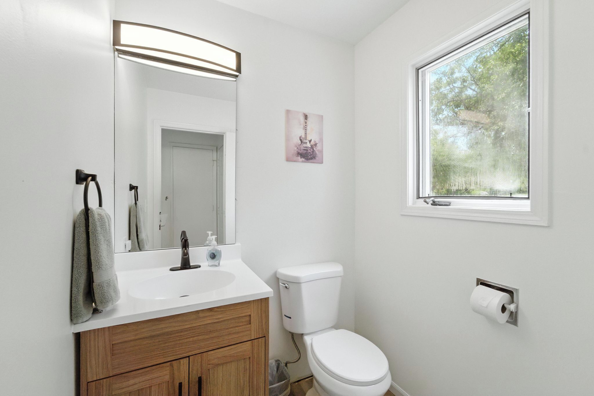 A bathroom with a white sink, wooden vanity, a toilet, a mirror, and a window with a view of trees.