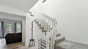 Una escalera con barandilla blanca y peldaños alfombrados, que conduce a un piso superior. Un estante de metal negro está apoyado contra la pared cerca de la escalera.