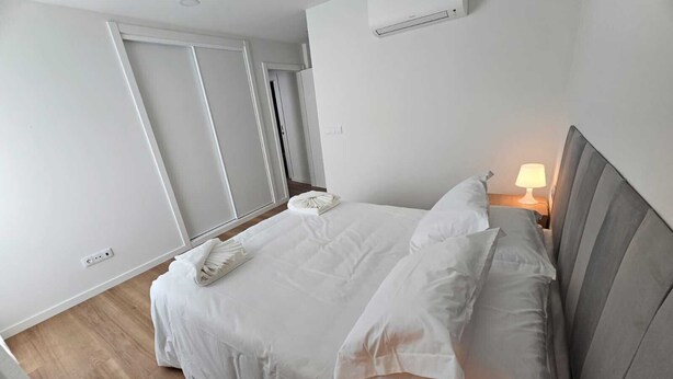 Una cama bien hecha con sábanas blancas, una lámpara de noche y un aire acondicionado de pared.