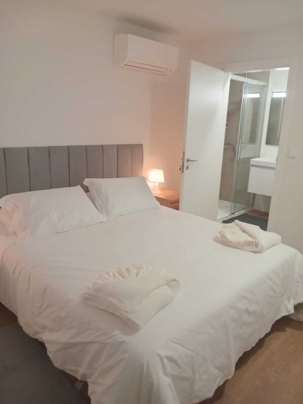 A neatly made bed with white linens and a folded towel, a bedside lamp, and a glass shower enclosure.