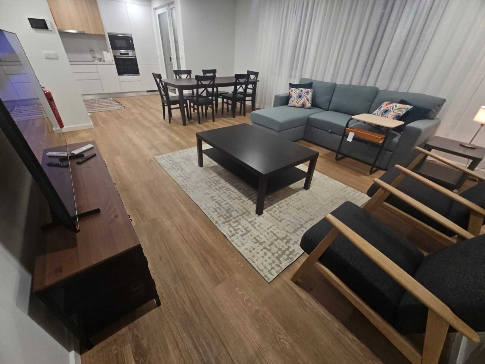 Een moderne woonkamer met een bank, salontafel en stoelen. In de achtergrond bevindt zich een keuken met kasten en apparatuur.