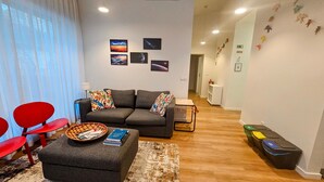 A living room with a grey sofa, red chairs, a small table, and a basket of magazines. There are framed pictures on the wall and a hallway with a white cabinet. - Sky Dream Apartment (Ponta Delgada)