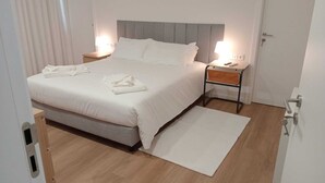 A neatly made bed with white linens and towels, a bedside table with a lamp, and a small rug on the wooden floor. - Sky Dream Apartment (Ponta Delgada)
