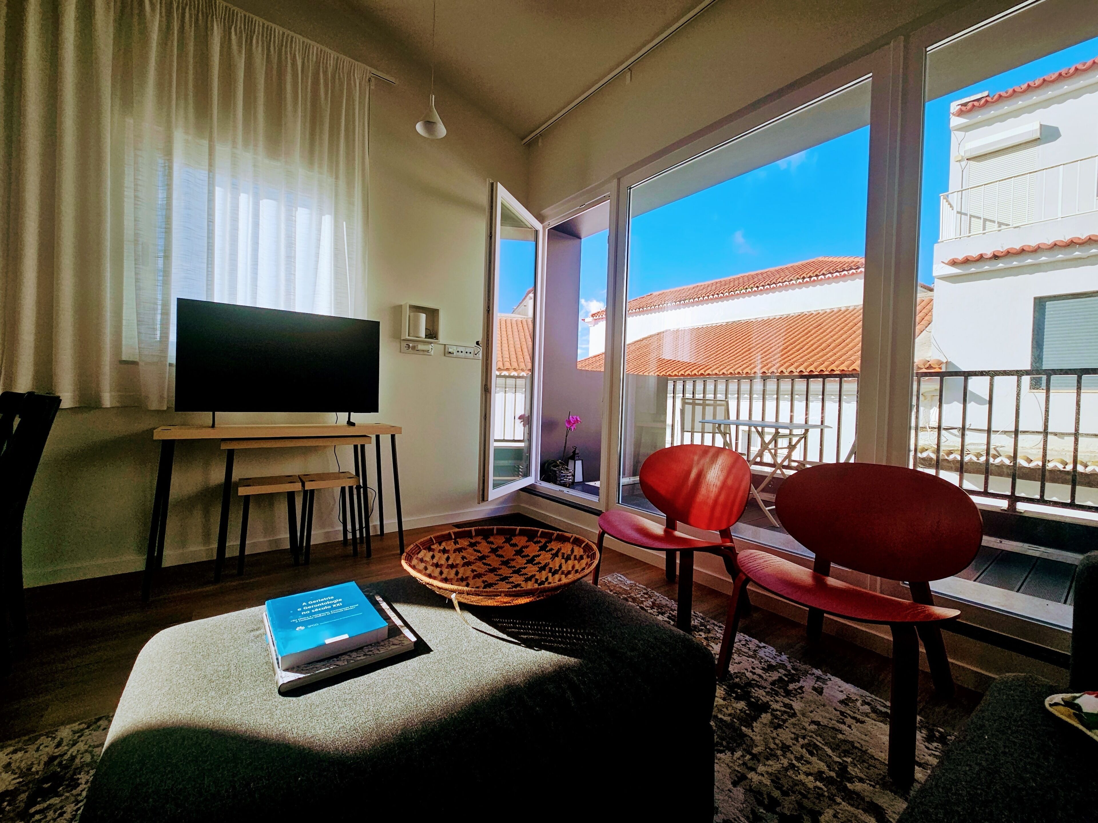 A living room with a TV, a sofa, a chair, a small table, and a window with a view of buildings.