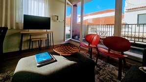 A living room with a TV, a sofa, a chair, a small table, and a window with a view of buildings. - Sky Dream Apartment (Ponta Delgada)